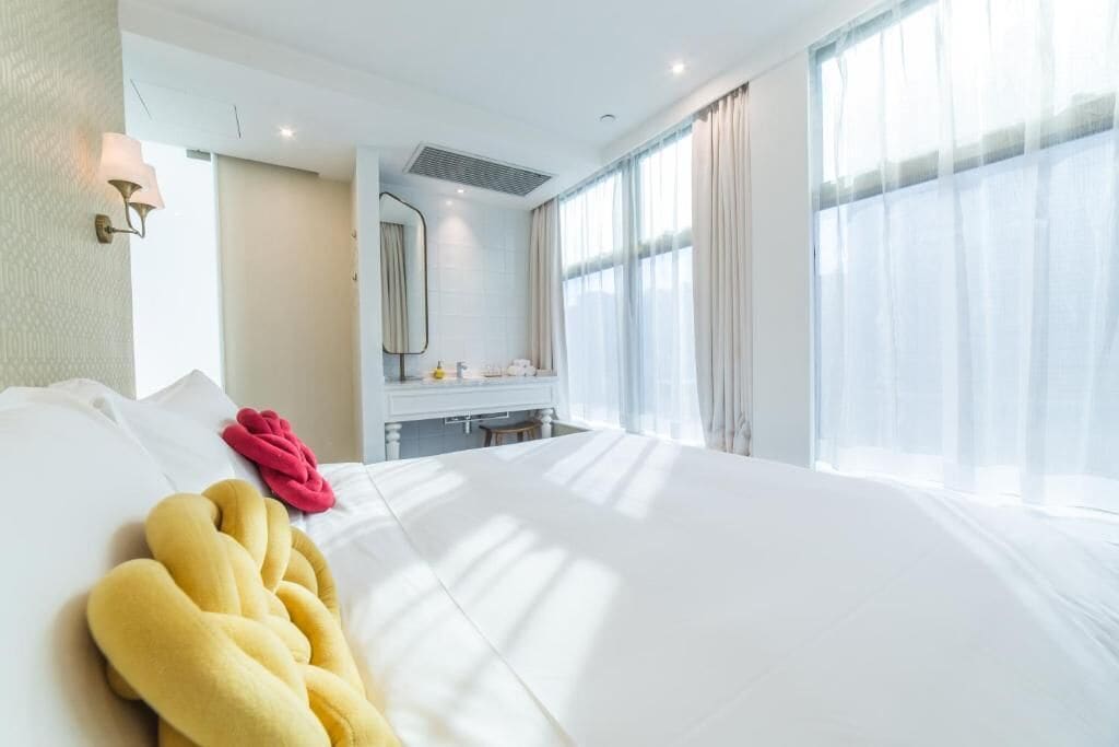 A hotel room with a large bed, a bedside table, a mirror, and a window with sheer curtains.
