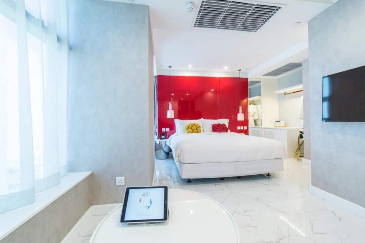 A modern hotel room with a large bed, a bedside table with a tablet, and a red accent wall.