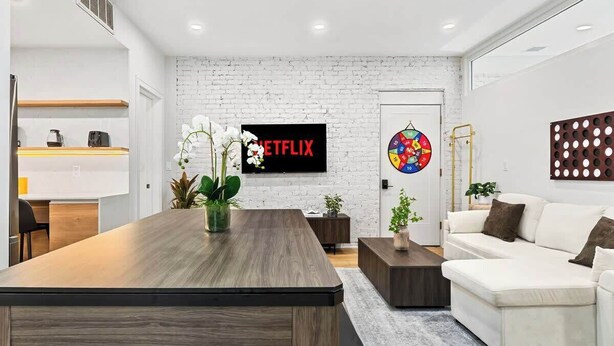 Un salón moderno con una mesa de comedor de madera, un sofá blanco y un televisor montado en una pared de ladrillo blanco.