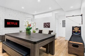 A modern living room with a wooden dining table, a sofa, a TV displaying 'NETFLIX,' and a pinball machine.