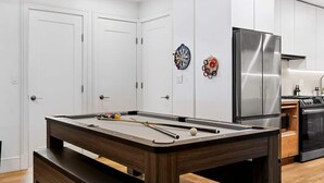 Una mesa de billar en una habitación de paredes blancas, un área de cocina con electrodomésticos de acero inoxidable y una puerta con un reloj en la pared.