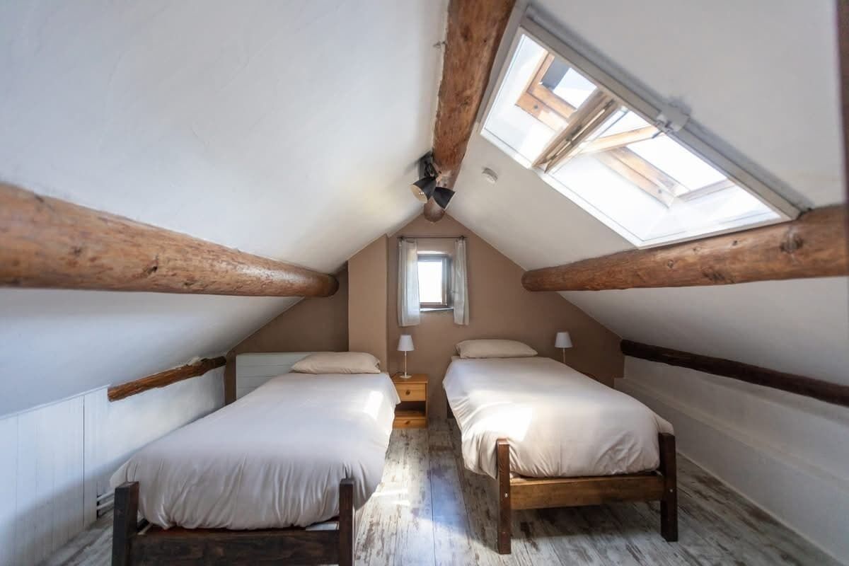 Une petite chambre mansardée avec deux lits en bois, une lucarne et une fenêtre.