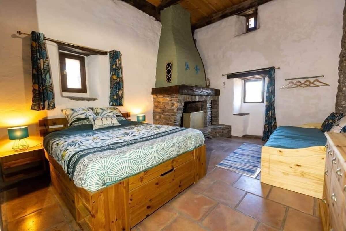 A rustic bedroom with a wooden bed, a stone fireplace, and a small window.
