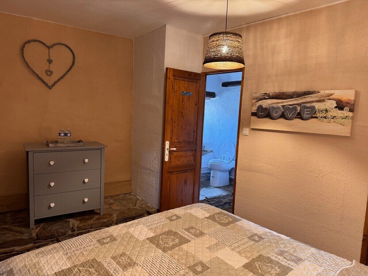 Une chambre à coucher avec un lit, une commode ornée de boutons en forme de cœur, une porte en bois et un cadre photo fixé au mur, sur lequel figure le mot « AMOUR ».