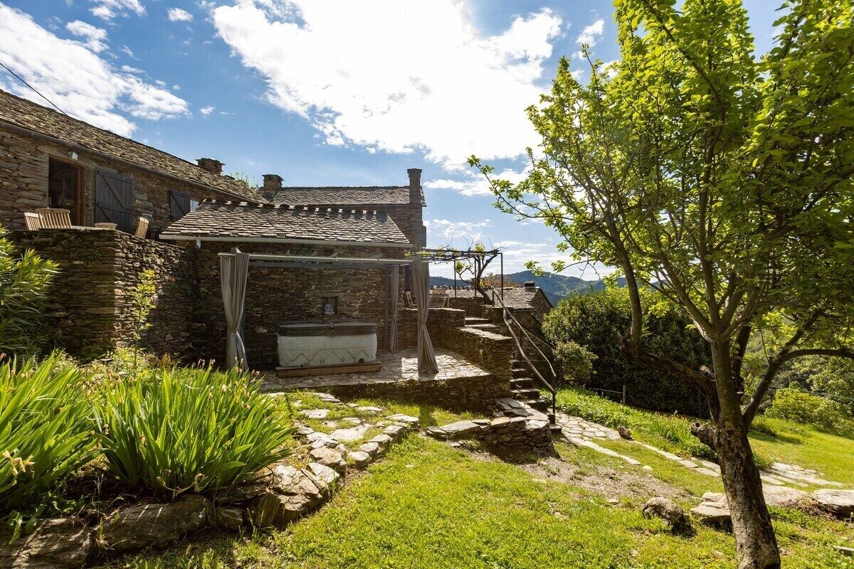 A rustic stone building with a covered outdoor seating area, stone steps leading up to a wooden deck, and a tree-lined path.