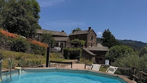 Una piscina azul cristalina con cubierta de madera, rodeada de edificios de piedra y abundante vegetación.