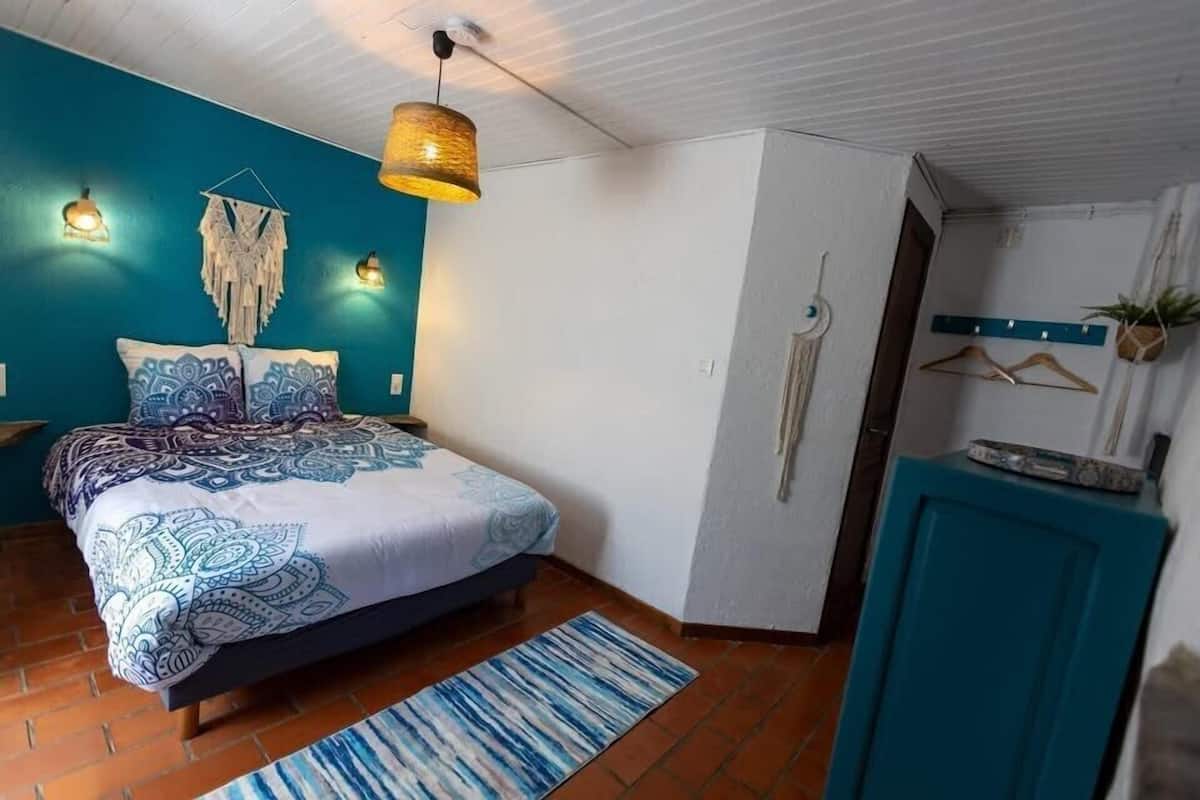 A bedroom with a bed, a blue wall, a hanging light fixture, and a small cabinet.