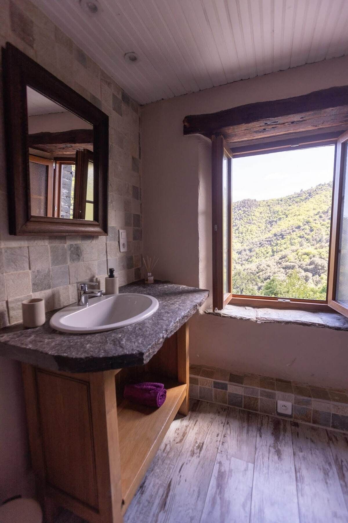 Une salle de bain avec un lavabo en pierre, un meuble-lavabo en bois, un grand miroir et une fenêtre donnant sur un paysage vallonné.