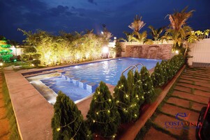 Piscina com cachoeira, cercada por vegetação iluminada e palmeiras.