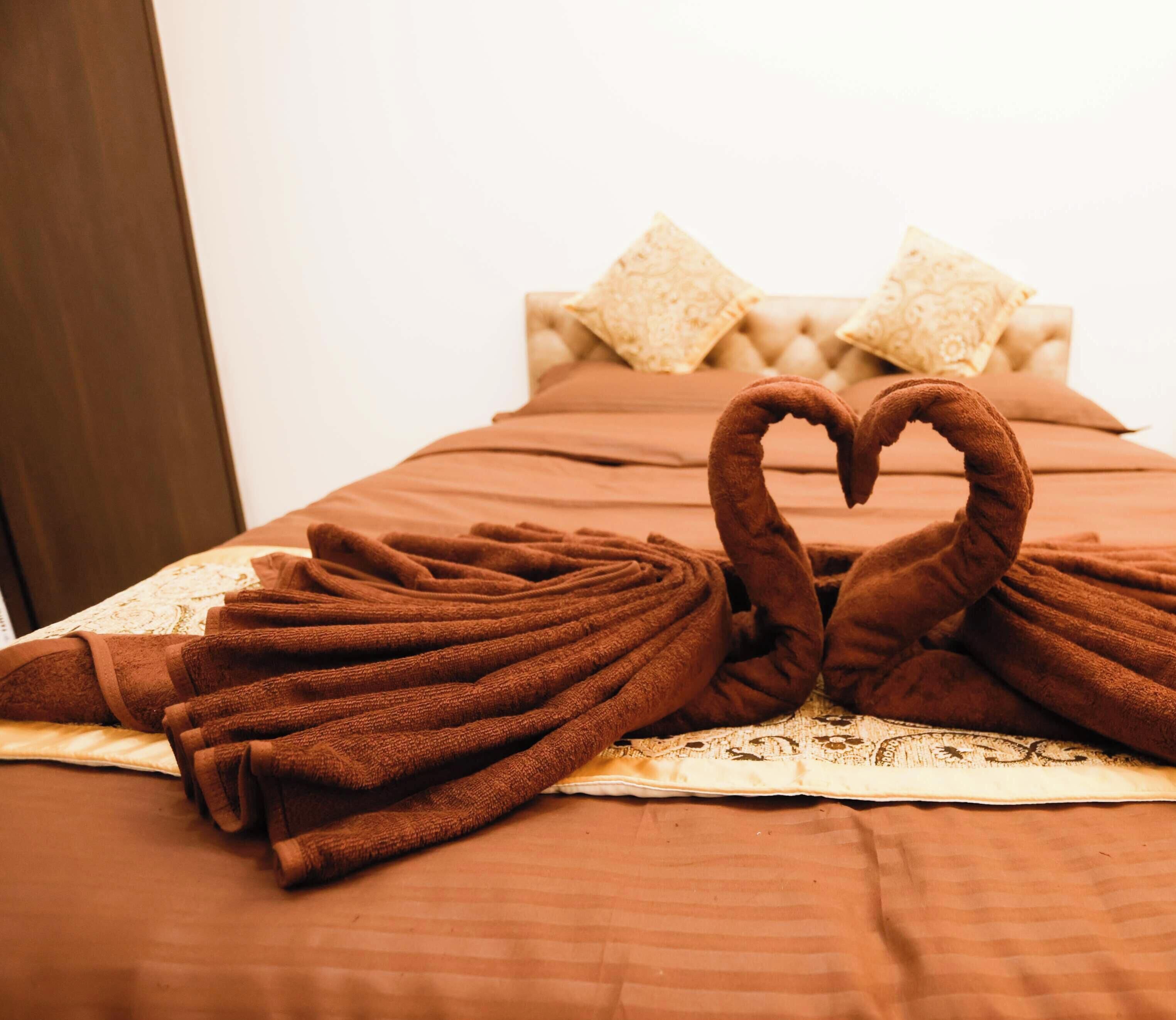 A bed with brown towels arranged in a heart shape, a lace bedspread, and matching pillows.