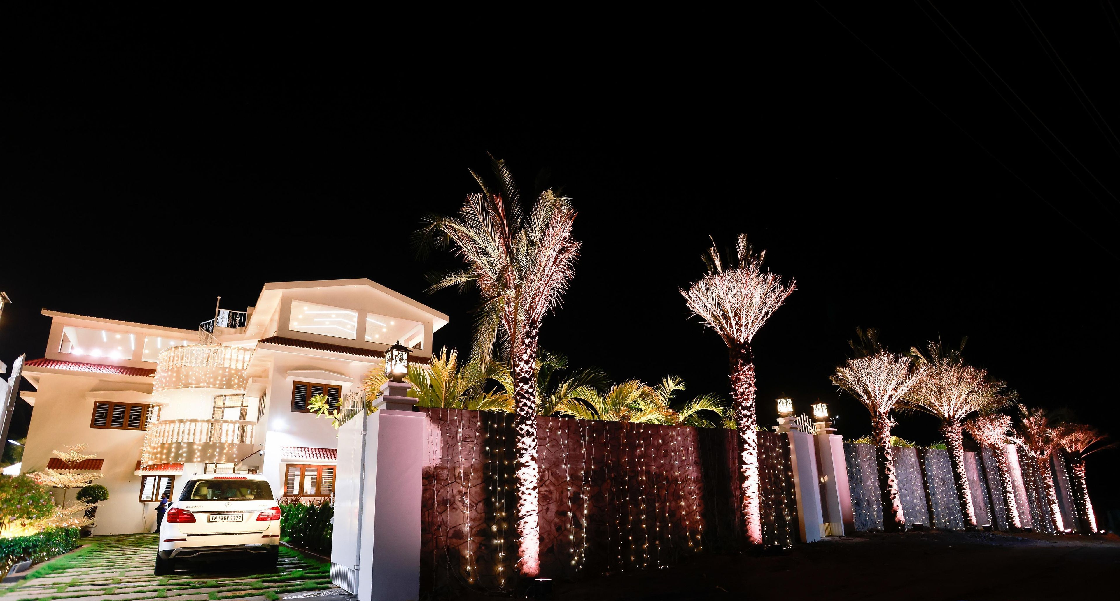 A modern house with a white car parked in the driveway, surrounded by well-lit palm trees and a decorative fence.