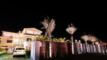A modern house with a white car parked in the driveway, surrounded by well-lit palm trees and a decorative fence.