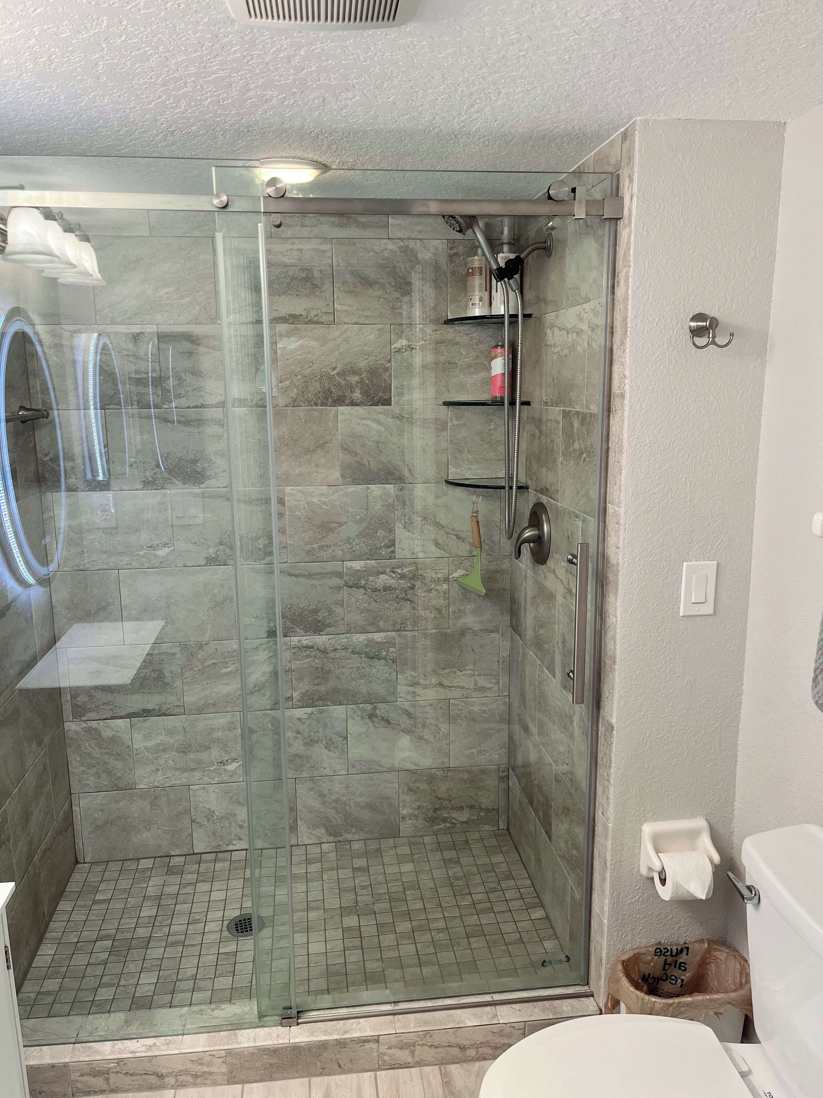 A modern bathroom with a glass-enclosed shower, marble tiles, a toilet, and a trash bin.