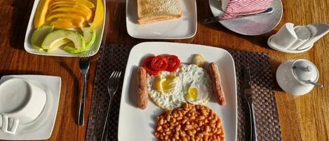Un desayuno con tostadas, huevos, salchichas, frijoles y fruta sobre una mesa de madera.