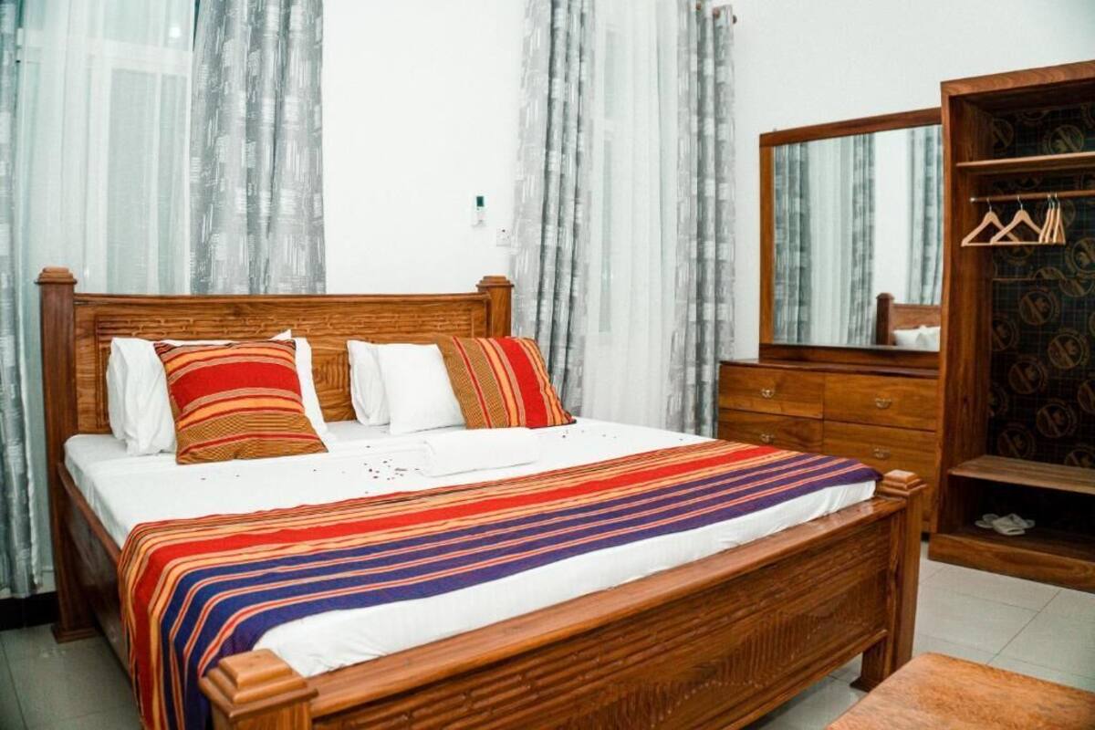 A wooden bed with a striped bedspread and pillows, a wooden dresser with a mirror, and a wardrobe with hangers.