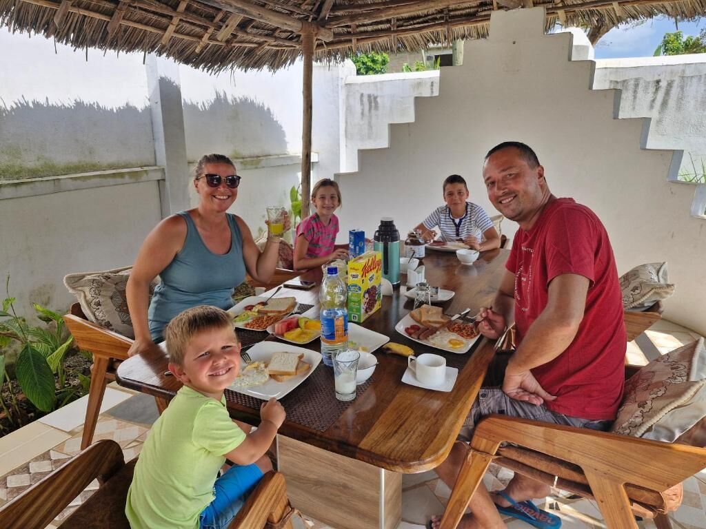 Una familia cenando en una mesa al aire libre bajo un techo de paja, rodeada de plantas y con vistas al paisaje.