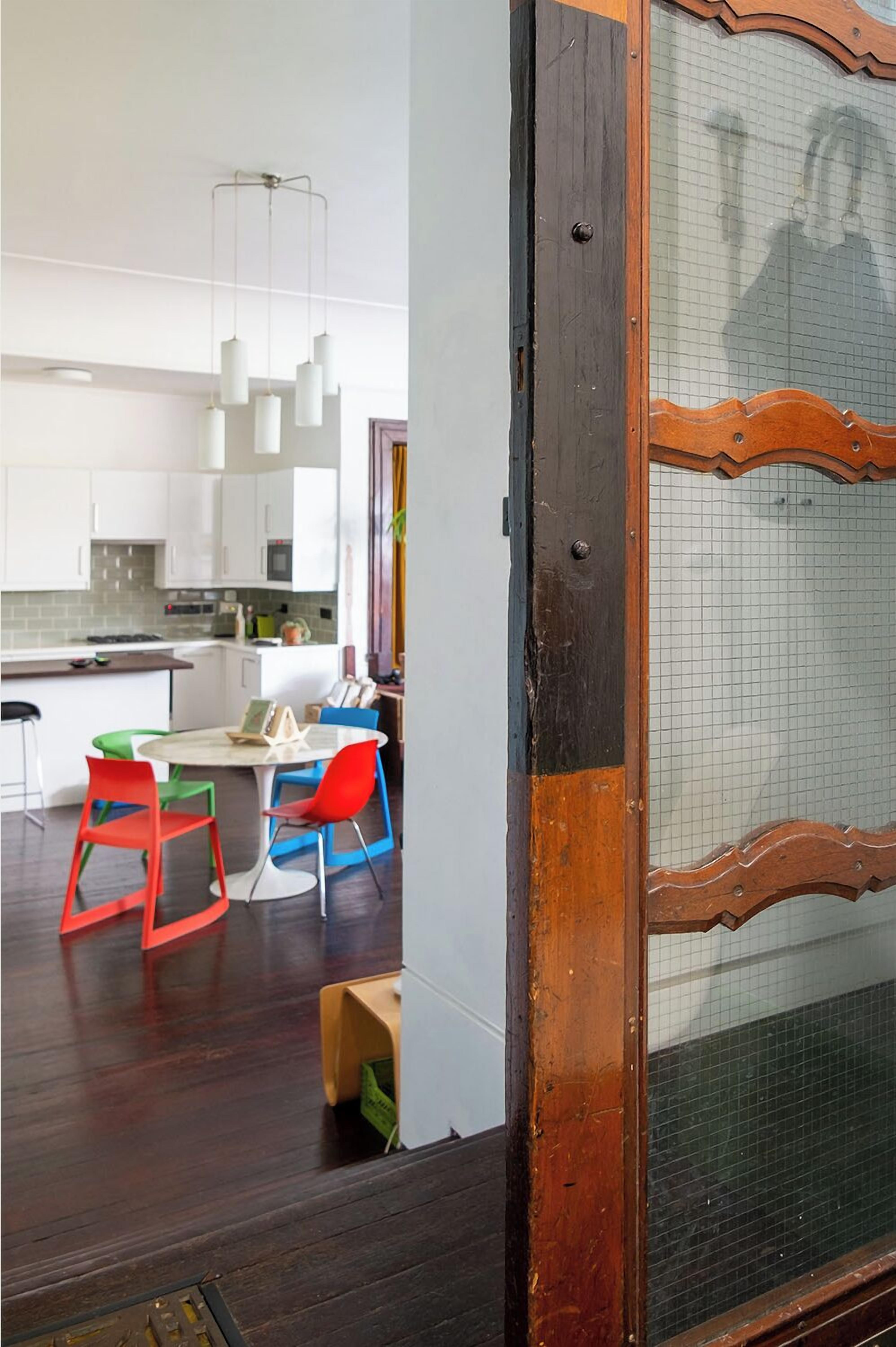 Una cocina moderna con armarios blancos, un comedor con sillas coloridas y una puerta de madera con mosquitera.