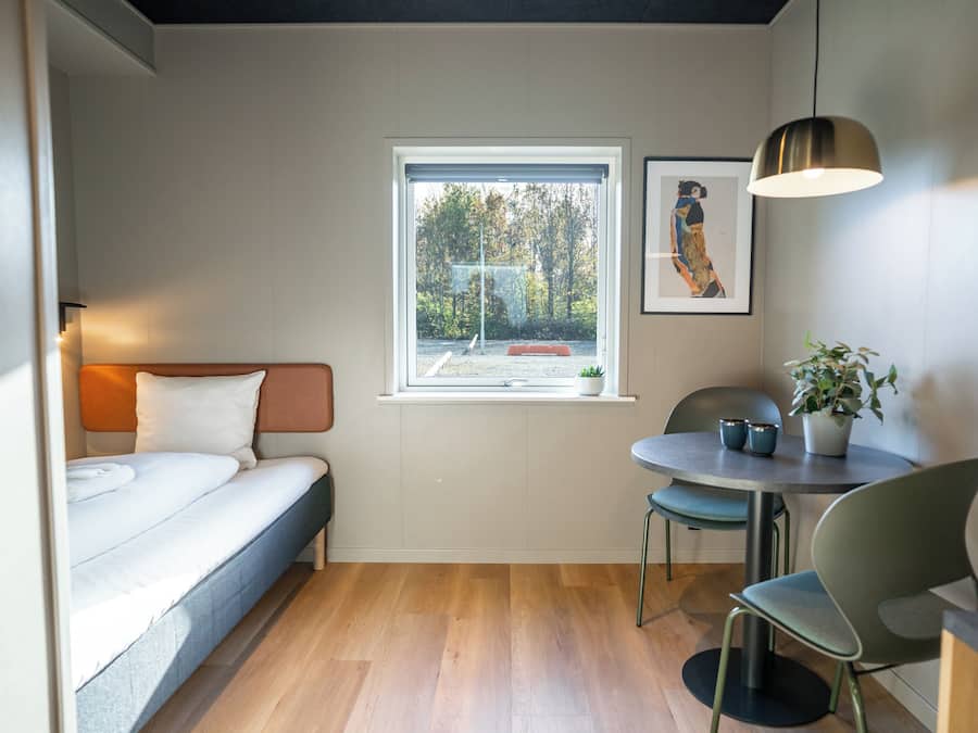 Habitación pequeña con cama, una mesita con sillas, ventana con vista a árboles y una lámpara colgante.