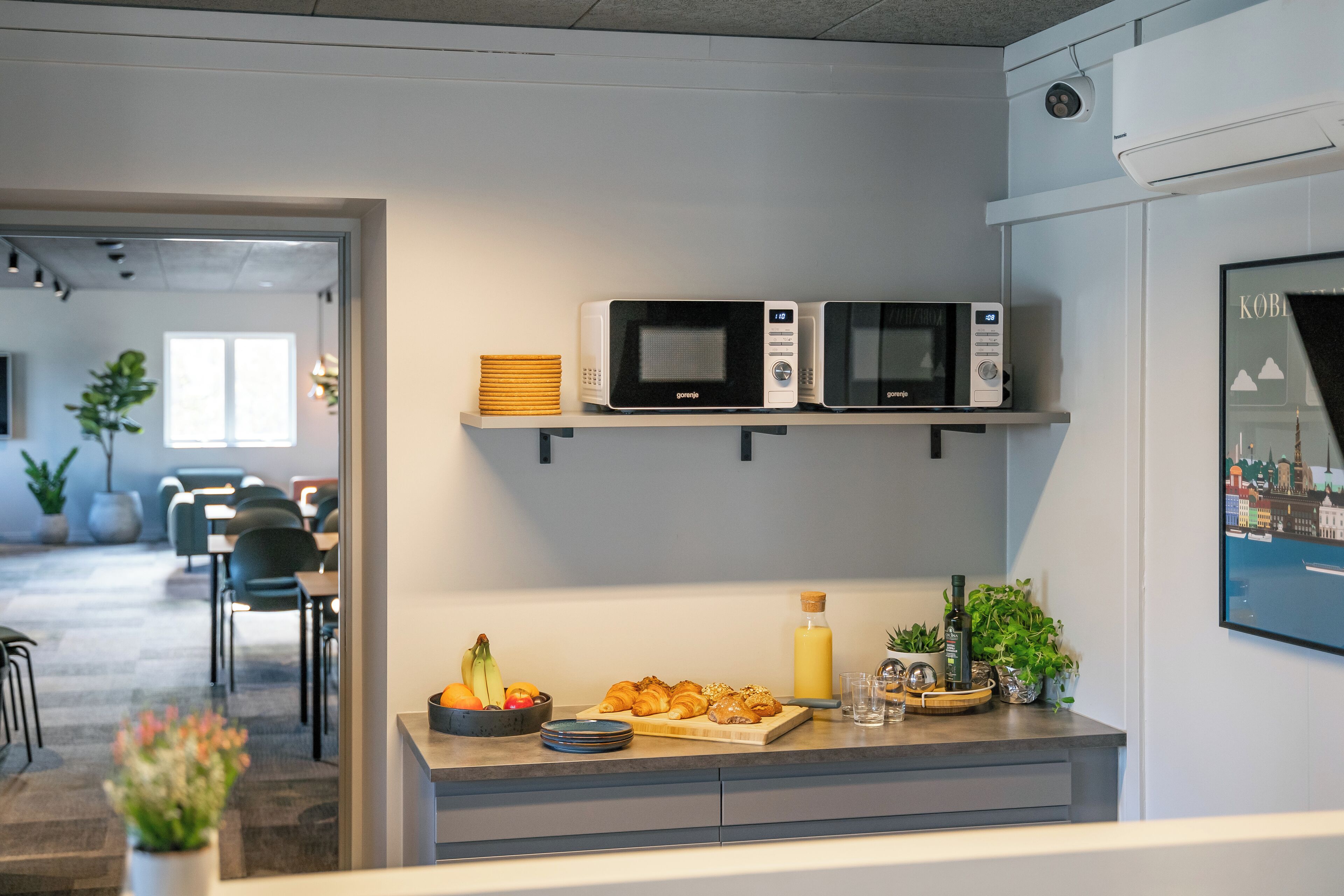 Ett modernt kök med två mikrovågsugnar på en hylla, en fruktskål, en skärbräda med croissanter och en flaska apelsinjuice.