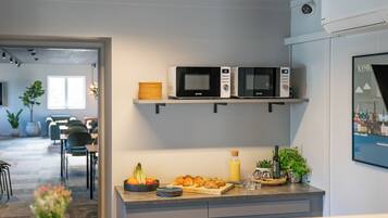 Ett modernt kök med två mikrovågsugnar på en hylla, en fruktskål, en skärbräda med croissanter och en flaska apelsinjuice.
