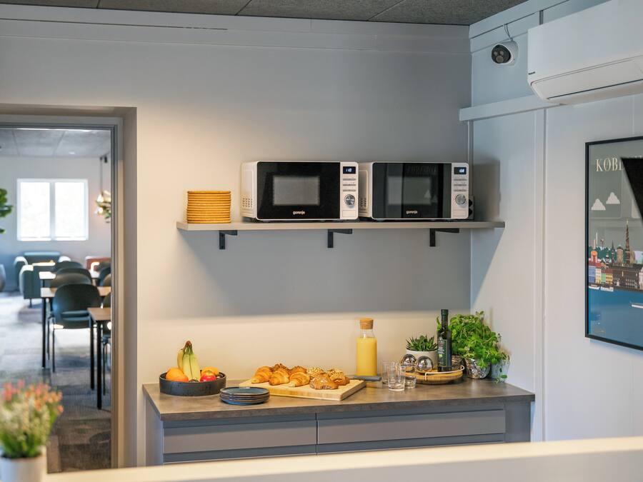 Ett modernt kök med två mikrovågsugnar på en hylla, en fruktskål, en skärbräda med croissanter och en flaska apelsinjuice.