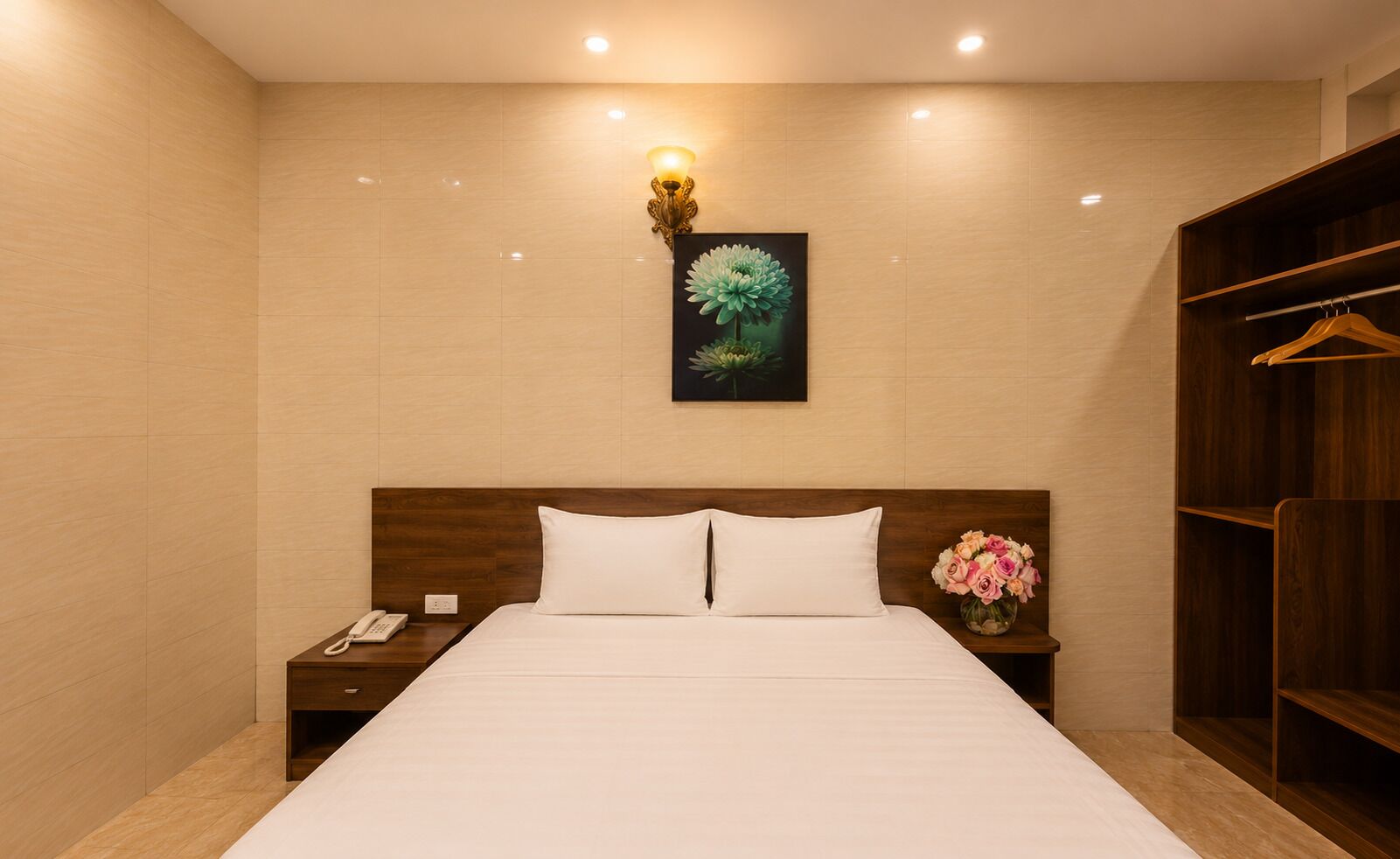 A hotel room with a large bed, wooden headboard, bedside tables, a phone, a vase with flowers, a wall-mounted light fixture, and a wardrobe.