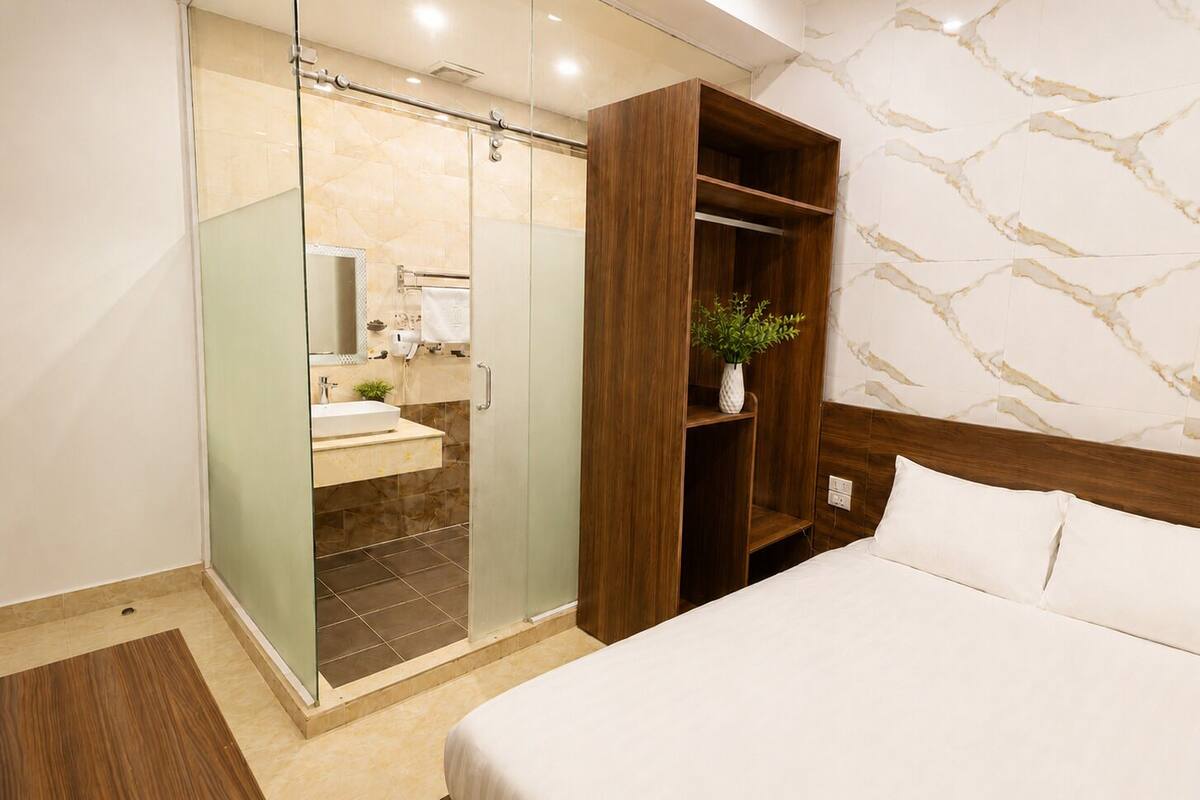 A hotel room with a bed, a wooden wardrobe, a glass shower enclosure, and a marble-patterned wall.