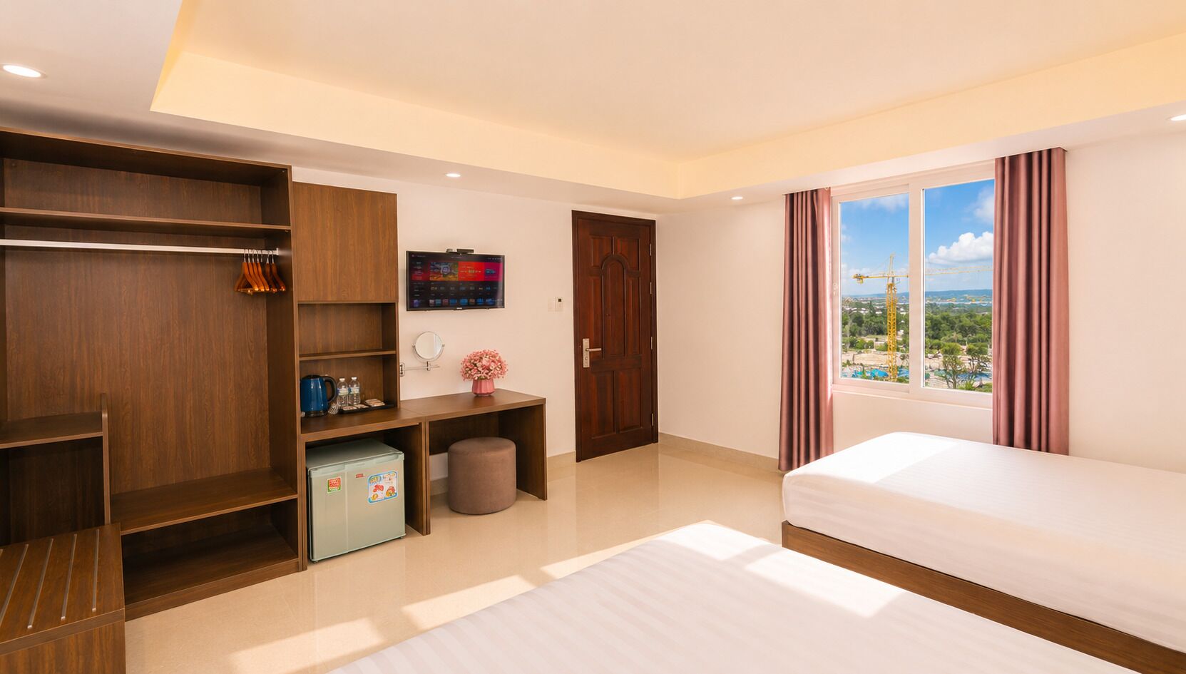 A hotel room with a bed, a wardrobe, a desk with a computer, a small fridge, a window with a view, and a curtain.