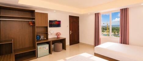 A hotel room with a bed, a wardrobe, a desk with a computer, a small fridge, a window with a view, and a curtain.