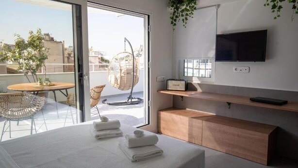 A modern hotel room with a large bed, a wooden bedside table, a TV mounted on the wall, and a balcony with wicker chairs and a hanging chair.