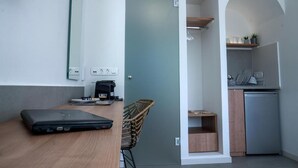 A compact room with a laptop on a wooden table, a wicker basket, a small kitchenette with a dishwasher, and a door leading to another area.