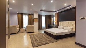 Una habitación de hotel moderna con una cama grande, una zona de estar con sofá y sillón, un escritorio y un ventanal con cortinas.