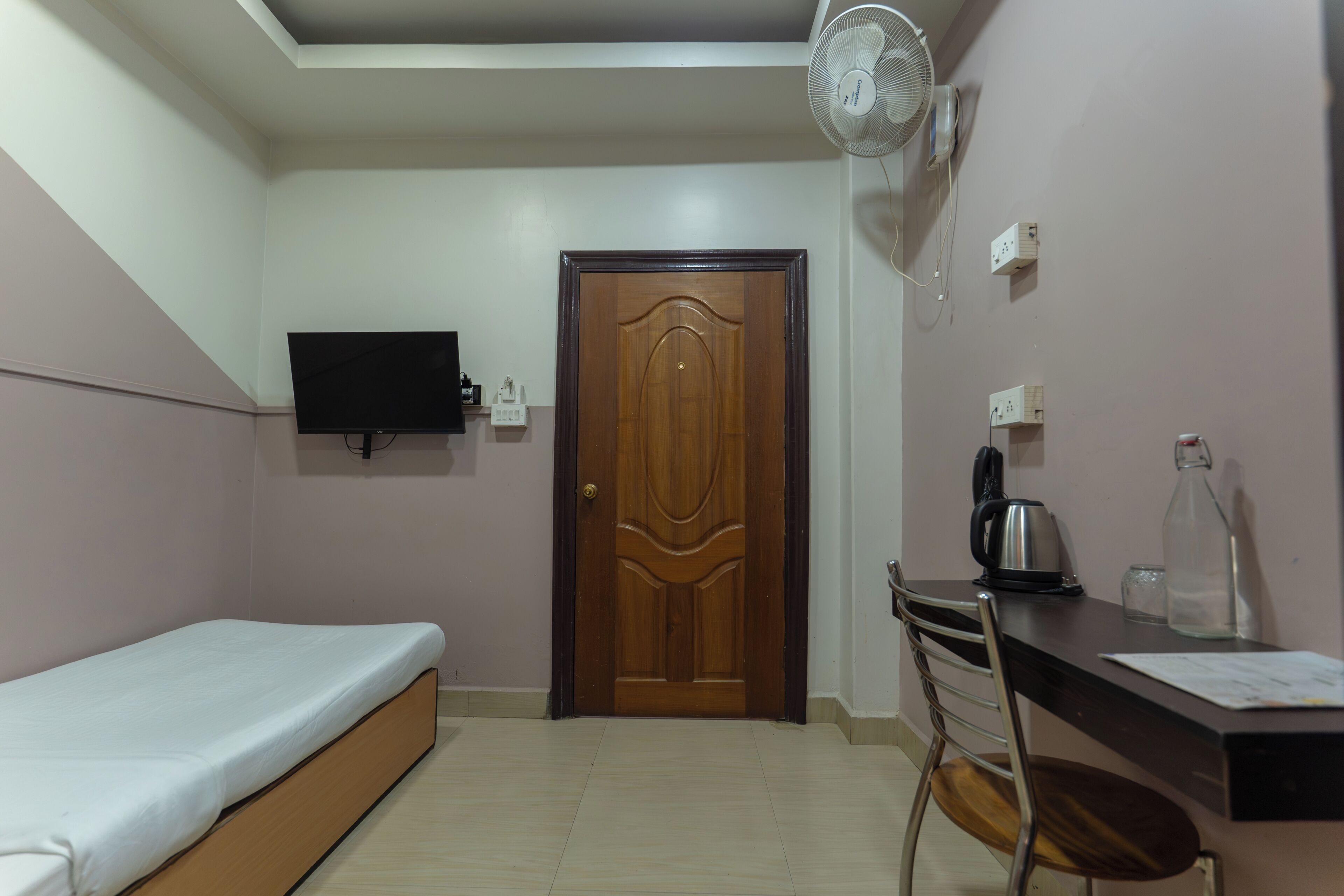 A room with a bed, a desk with a chair, a television, a wooden door, and a ceiling fan.
