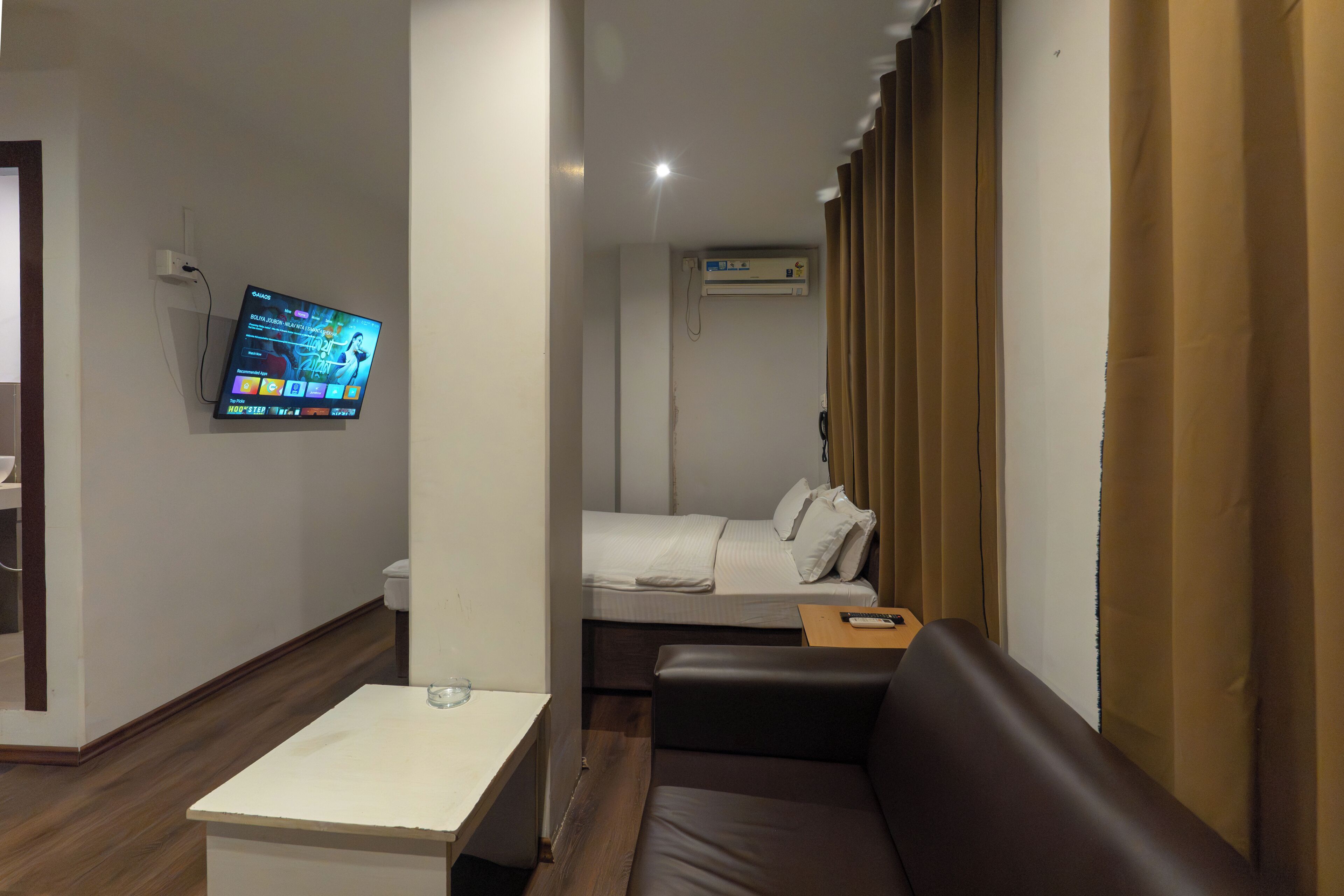 A hotel room with a bed, a sofa, a TV mounted on the wall, and an air conditioning unit.