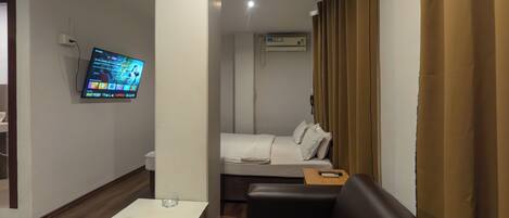 A hotel room with a bed, a sofa, a TV mounted on the wall, and an air conditioning unit.
