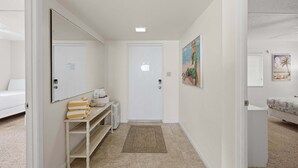 Un pasillo con una puerta blanca, un banco con toallas, una mesita con objetos y un cuadro en la pared.
