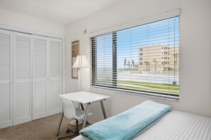 A room with a bed, a desk, a chair, a window with blinds, and a door with louvered shutters.