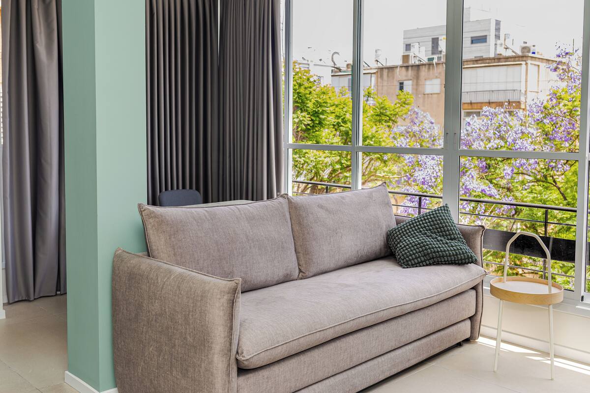 A modern living room with a grey sofa, a small round wooden table, and a view of buildings and trees through large windows.