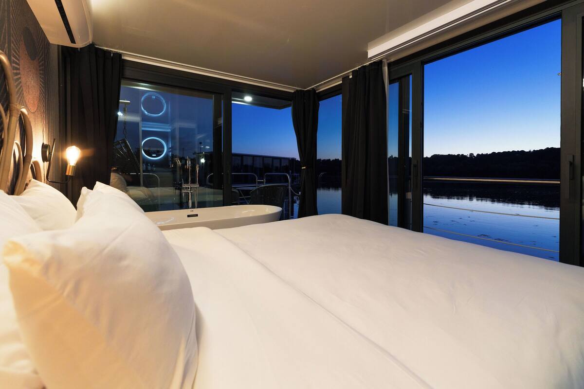 Een moderne hotelkamer met een groot bed, een badkamer met een ronde spiegel en uitzicht op een waterpartij in de schemering.