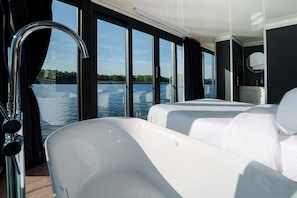 Un intérieur moderne avec deux lits blancs, une grande fenêtre donnant sur un lac, et une salle de bain avec un miroir et un lavabo.