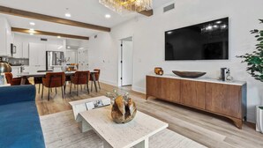 Una sala de estar moderna con comedor, piso de madera, una mesa de centro blanca con un adorno decorativo, un televisor de pantalla plana grande montado en la pared y un mueble de madera con un tazón encima.