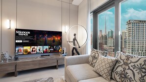 A modern living room with a large flat-screen TV displaying 'Stranger Things', a sofa with patterned cushions, a coffee table, and a cityscape view through large windows.
