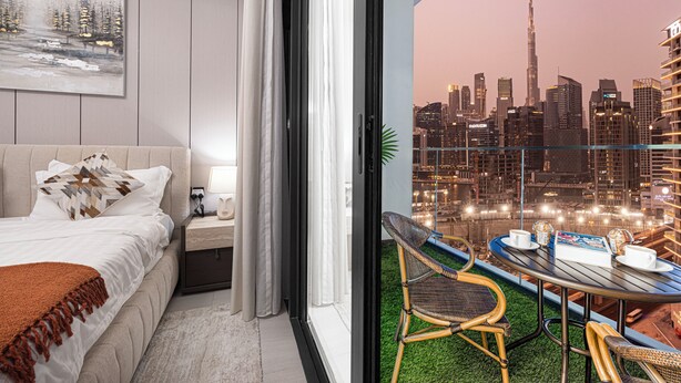 A hotel room with a bed, nightstand, and a balcony with a table and chairs. The balcony overlooks a cityscape with tall buildings.