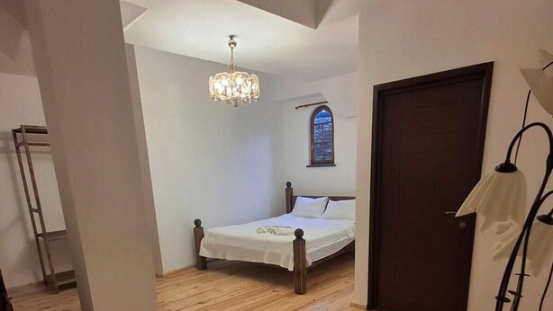 Quarto com piso de madeira, cama com roupa de cama branca, um candelabro, uma janela e uma porta.
