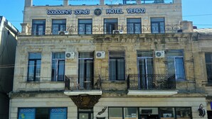 Um edifício de dois andares com a placa do Hotel Verdi. Possui várias janelas, algumas com aparelhos de ar condicionado, e varandas com guarda-corpos de metal.