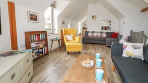 A spacious living area with a high ceiling, a yellow armchair, a grey sofa, and a wooden coffee table with pastries. - Coastguard Lookout (Weymouth)