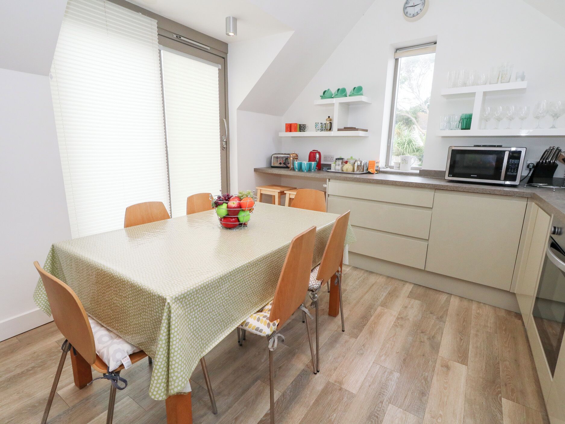 Un coin repas avec une table dressée pour quatre personnes, une cuisine équipée d’un four à micro-ondes et d’ustensiles, et une fenêtre avec des stores.