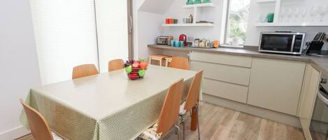 Un coin repas avec une table dressée pour quatre personnes, une cuisine équipée d’un four à micro-ondes et d’ustensiles, et une fenêtre avec des stores.