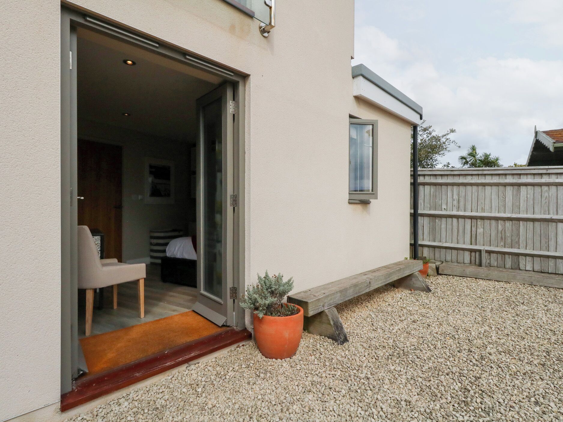 A residential building with a sliding glass door leading into a room with a bed and a chair. There is a small window and a wooden bench outside.