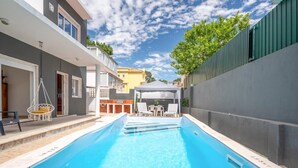 En swimmingpool med krystalklart, blåt vand, omgivet af en moderne bygning med en grå facade, en hvid balkon og et grønt hegn.